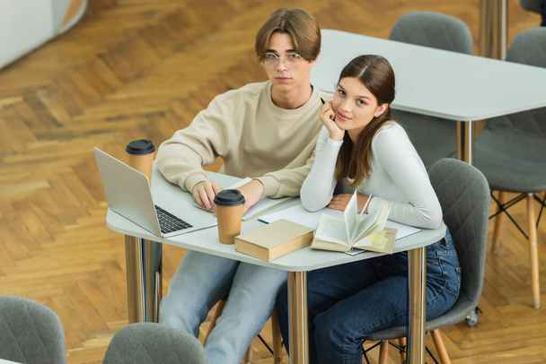 smiling teenage girl looking at camera near friend in eyeglasses and laptop in library - Fotografie, Obrázek