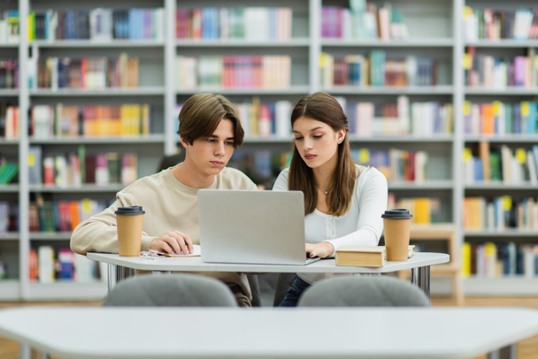 teenage friends looking at laptop while sitting in library reading room - Fotó, kép