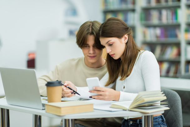 подростки смотрят на смартфон возле книг и ноутбука в читальном зале - Фото, изображение