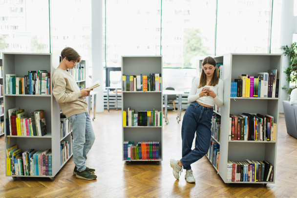 teen students with book and mobile phone standing near shelves in library - Photo, image