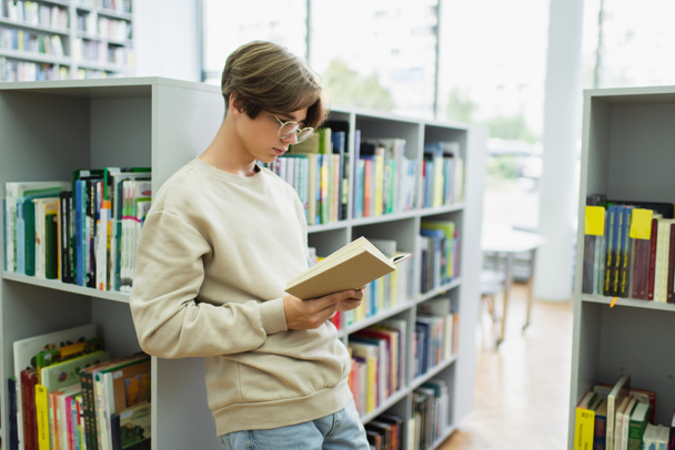 student in eyeglasses reading book while standing near shelves in library - Foto, imagen