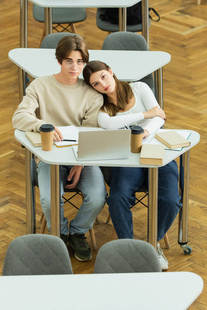 teenage couple looking at camera near laptop and books in library reading room - Valokuva, kuva