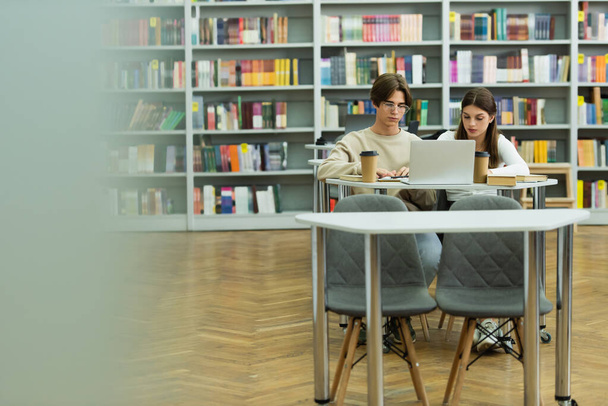 couple of teenagers sitting near laptop and paper cups in library on blurred foreground - Foto, afbeelding