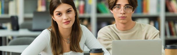 teenage girl smiling at camera near friend and laptop in blurred library, banner - Foto, immagini