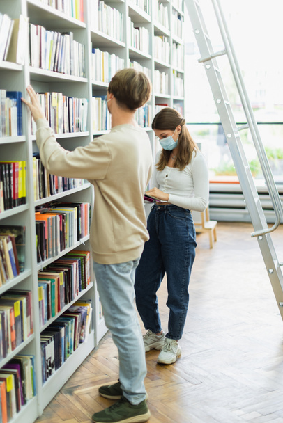 друзі-підлітки в медичних масках біля книжкових полиць і сходів в бібліотеці
 - Фото, зображення