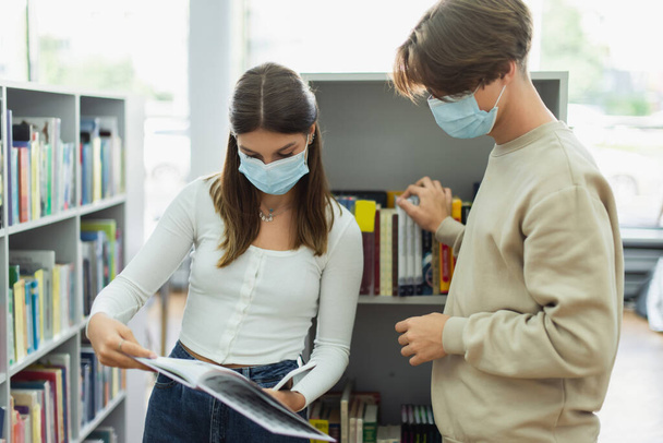 teenage girl in medical mask showing book to guy near racks in library - Fotó, kép