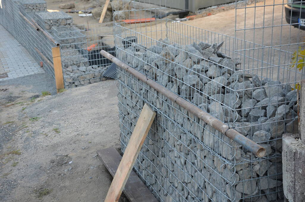 Bau einer Gabionen-Stützmauer als Teil des Hauszaunes. Arbeiter legen Geotextilien und darauf Drahtkörbe, die aus Granitsteinen bestehen. Anspruchsvolle Handarbeit - Foto, Bild