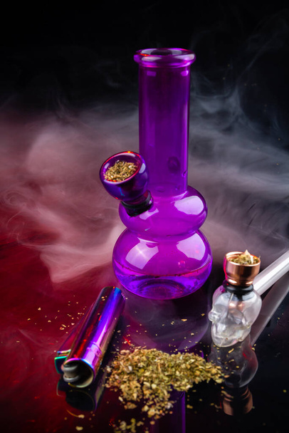 Vertical shot of glass bong with smoking pipe and pile of weed. Set of marijuana and smoking devices. - Foto, imagen