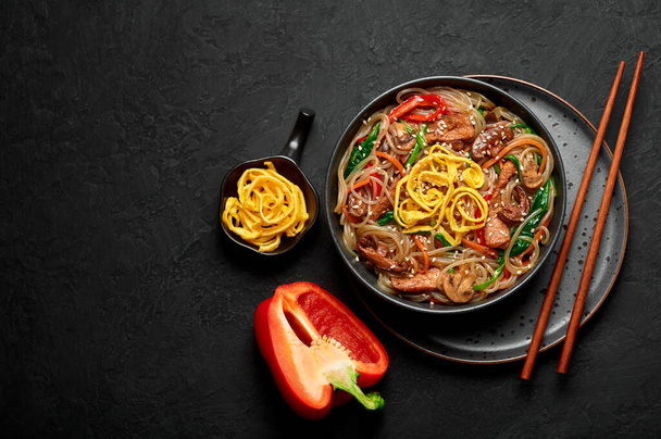 Japchae in black bowl on dark slate table top. Korean cuisine glass chapchae noodles dish with vegetables and meat. Asian traditional food. Authentic meal. Top view. Copy space - Foto, Imagen