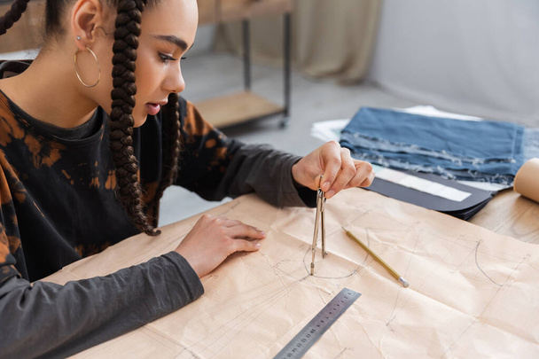 African american designer drawing with compass on sewing print near blurred cloth in workshop  - Foto, afbeelding