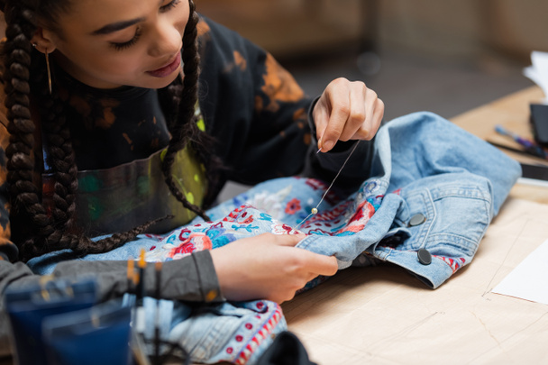 African american designer decorating denim jacket with embroidery in workshop  - Foto, afbeelding
