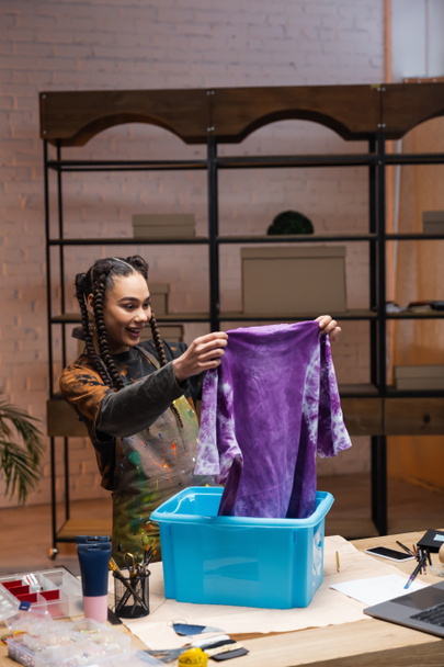 Smiling african americn designer holding t-shirt near container in workshop  - Foto, Imagem