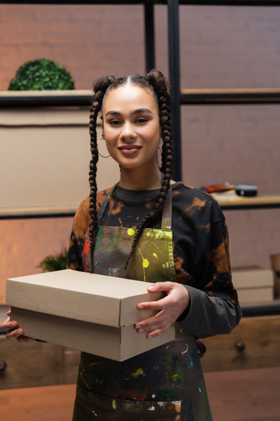 Smiling african american designer holding carton box in workshop  - 写真・画像