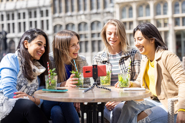 Τέσσερις πολυεθνικές φίλες, τουρίστες ή φοιτητές που κάθονται σε μια βεράντα καφέ στο κέντρο της πόλης χρησιμοποιώντας το κινητό τηλέφωνο για μια βιντεοκλήση. Υψηλής ποιότητας φωτογραφία - Φωτογραφία, εικόνα