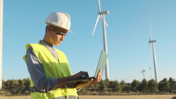 Female engineer using a laptop while working outdoors at a wind turbine farm. Sustainable lifestyles, renewable energy and technology concept. - Filmagem, Vídeo