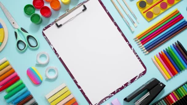 Colorful school supplies placed on blue background with white plain paper in the middle. Back to school concept. Top view, flat lay - Footage, Video