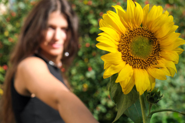 Young cute girl with long straight hair in a black crop top with a sunflower focus on a flower - Foto, Imagem