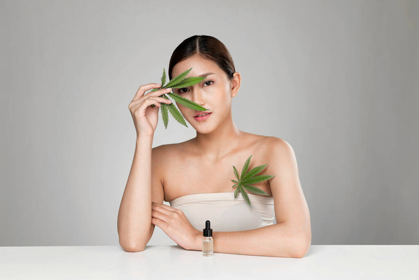 Portrait of young ardent woman with healthy fresh skin holding green hemp leaf and cbd oil. Combination of beauty and cannabis concept. - Foto, Imagen