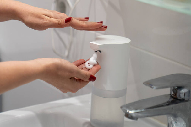 automatic sensor touchless soap dispenser, close up woman washing hands with soap and water, white foam, in the bathroom, under tap in the sink, personal hygiene and care, coronavirus prevention - Foto, Imagen