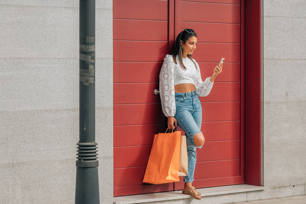 jovem menina latina na rua com sacos de compras e telefone celular - Foto, Imagem
