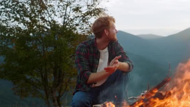Smiling traveler warm fire on camp. Joyful millennial rest in mountains forest closeup. Happy guy warming hands by bonfire outdoors. Redhead tourist enjoy peaceful evening. Healthy lifestyle concept. - Metraje, vídeo