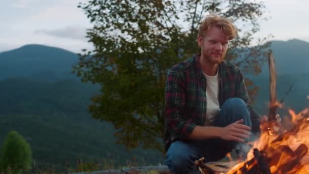 Active traveler camp forest in mountains. Close up redhead hiker sit bonfire on nature evening sunset. Cheerful camper throw log wood in fire. Young tourist spend weekend. Leisure relax travel concept - Footage, Video