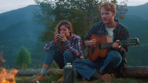 Wanderlust couple spend weekend in mountains. Young family enjoy camp evening outdoors closeup. Thoughtful travelers play music instrument guitar by bonfire. Tourism youth lifestyle leisure concept. - Imágenes, Vídeo