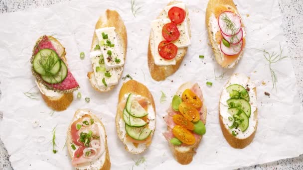 Variety of small sandwiches with cream cheese, vegetables and salami. Made with cream cheese, cucumber, radish, tomatoes, salami, chives on a white baking paper. Top view. Flat lay - Video