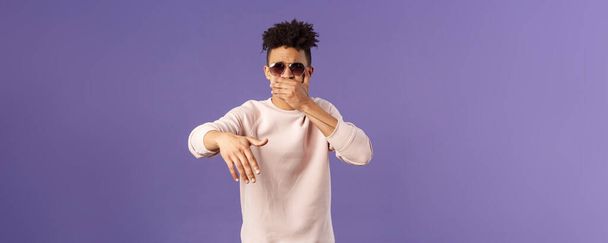 Waist-up portrait of cool and sassy, young carefree guy with dreads and sunglasses, cover mouth to beatbox, waving hand in rhythm music, singing rap or attend hip-hop party, purple background. - Photo, Image