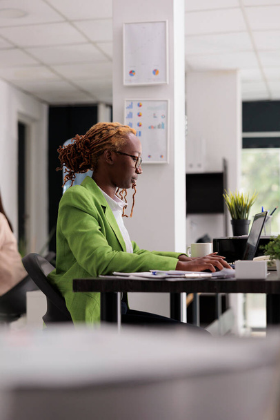 Νεαρή σοβαρή γυναίκα δακτυλογραφεί σε φορητό υπολογιστή στο γραφείο συνεργασίας, επαγγελματική εταιρική διευθυντής κάθεται στο γραφείο στο χώρο εργασίας. Εστιασμένη Αφροαμερικανός εργαζόμενος που εργάζεται στον υπολογιστή, ημέρα εργασίας των εργαζομένων - Φωτογραφία, εικόνα