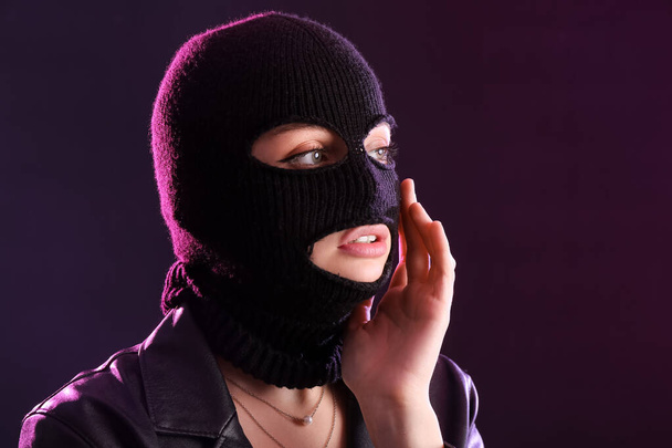Young woman in balaclava and leather jacket with hand near face against dark background - Valokuva, kuva