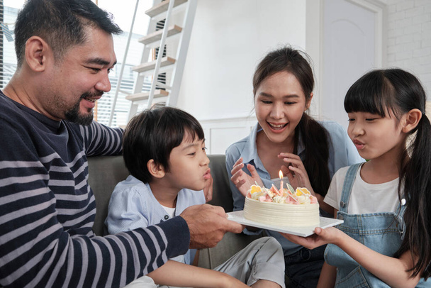 Happy Asian Thai family, young son is surprised with birthday cake, blows out candle, and celebrates joy party with parents and sister in living room together, wellbeing domestic home special event. - Foto, Imagen