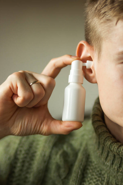 Ο παντρεμένος καυκάσιος χρησιμοποιεί σπρέι για τα αυτιά. Mans χέρι κατέχει ένα λευκό μπουκάλι ψεκασμού αυτί με ακροφύσιο εγκατασταθεί στην τρύπα του αυτιού. Καθημερινή υγιεινή και καθαρίστε τα αυτιά από το κερί αυτιών. Έννοια υγειονομικής περίθαλψης. - Φωτογραφία, εικόνα