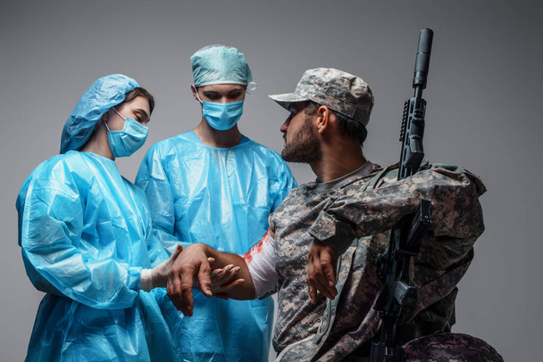 Photo of surgeon and his assistant treating and bandaging wounded soldier with rifle. - 写真・画像