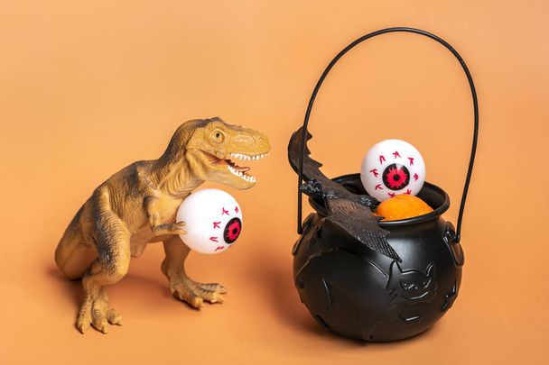 Toy dinosaur Tyrannosaurus holding eye in its paws, bowl with bat, pumpkin isolated on orange background Holiday greeting card Happy Halloween day creative minimal concept Calendar date 31 October. - Zdjęcie, obraz