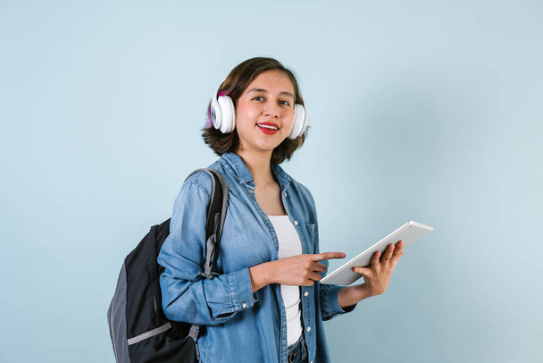 Young Hispanic student woman listen music with headphones and holding digital tablet over isolated blue background - Foto, imagen