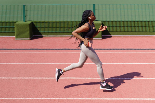 Mujer afroamericana disfruta corriendo todos los días en la pista roja escuchando música en los auriculares en el caluroso día de verano. Mujer joven trata de mantener un estilo de vida saludable en el campo de deportes del complejo residencial - Foto, imagen