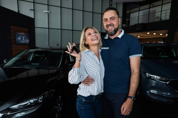 Happy caucasian couple hugging and woman holding new car keys in car showroom - Photo, Image