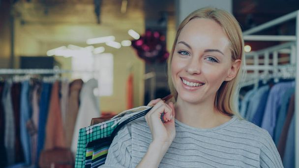Close-up portrait of cute blond girl standing with paper bags in clothing boutique, moving slightly, laughing and smiling happily and looking at camera. Stylish clothes is in background. - Φωτογραφία, εικόνα
