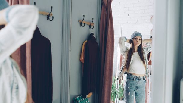 Mirror shot of young girl trying on clothes in fitting room. She is wearing denim jacket, jeans and hat, smoothing her hair, moving and looking in mirror to check image. Teenager shopping concept. - Foto, Imagen