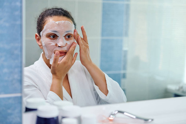 Charming dark-haired multi-ethnic woman in white bathrobe, looking at mirror in bathroom while applying moisturizing smoothing tissue mask on her face, taking care of the beauty and health of her skin - Fotó, kép
