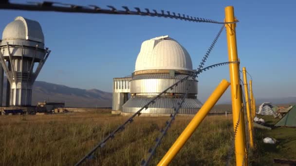 A large observatory is protected by a barbed fence. There is a long sharp wire around perimeter. Buildings with domes and a telescope are visible in distance. Yellow-green grass. Mountainous terrain - Footage, Video