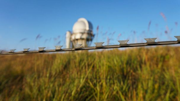 A large observatory is protected by a barbed fence. There is a long sharp wire around perimeter. Buildings with domes and a telescope are visible in distance. Yellow-green grass. Mountainous terrain - Valokuva, kuva