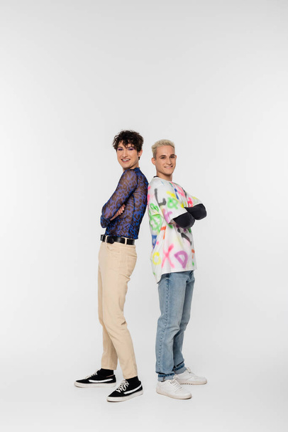 full length of cheerful gay man and queer person standing back to back with crossed arms on grey background - Fotó, kép