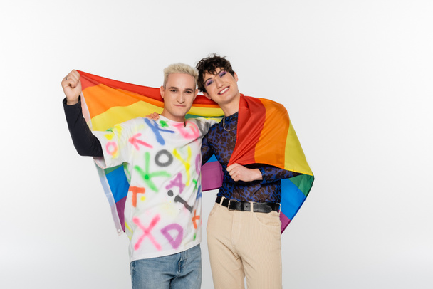 young and stylish gay man and queer person with lgbt flag smiling at camera isolated on grey - Foto, afbeelding