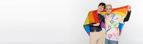 trendy queer person and blonde gay man with lgbtq flag smiling at camera isolated on grey, banner - Foto, imagen