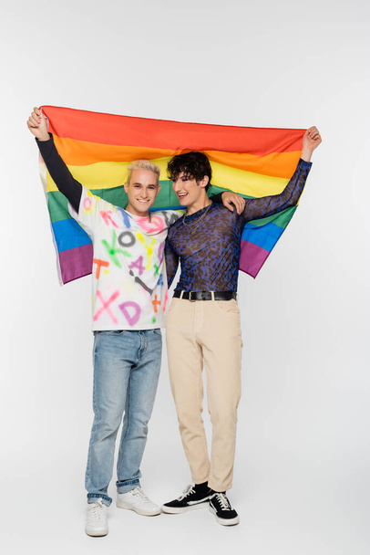 full length of gay man and nonbinary person holding lgbtq flag on grey background - Foto, imagen