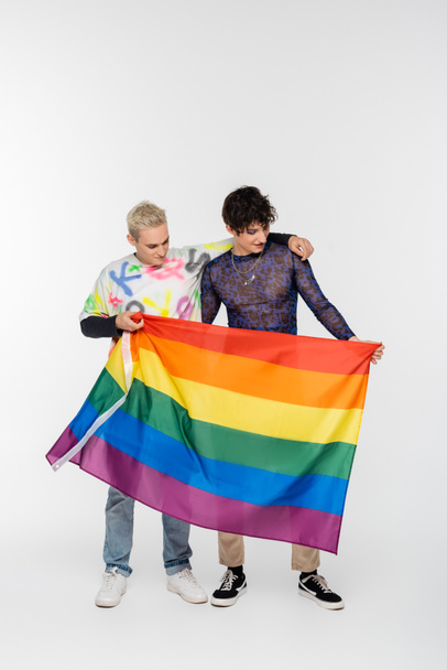 blonde gay man and brunette nonbinary person standing with lgbtq flag on grey background - Foto, immagini