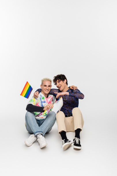 smiling gay man with small lgbtq flags sitting near queer person pointing with finger on grey background - Fotografie, Obrázek