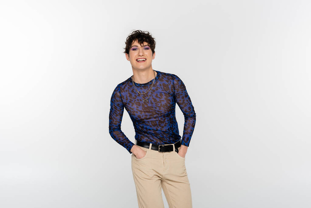 smiling nonbinary person standing with hands in pockets of beige trousers isolated on grey - Photo, image